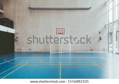 Front view of the court in the gymnasium hall; an indoor modern office stadium with a basketball basket and hoop, football goal, blue floor, a concrete wall with an undeployed projection screen above
