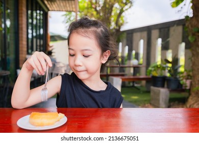 Front View Of Cute 5 Years Old Asian Girl Is Eating Delicious Sweet (donut) Outside The Cafe Shows Concept Of Happy Eating For Kid Or Child With Face Expression Of Enjoyment And Cheerfulness.