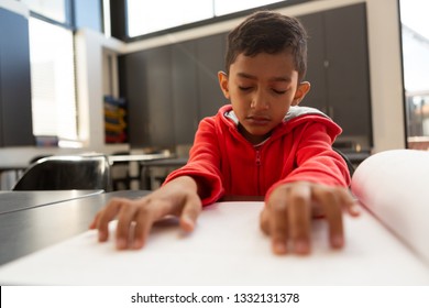 Front view of cute blind mixed-race schoolboy reading a braille book at desk in a classroom at elementary school
