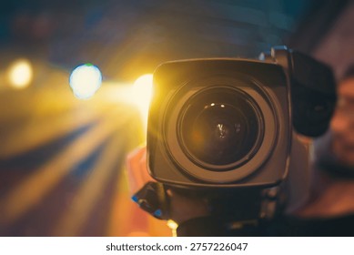 Front view of a digital cinema camera on a film set with golden bokeh and stage lights. Professional broadcasting equipment in dark studio