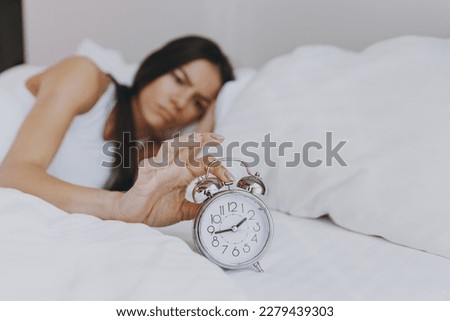 Front view distempered displeased irritated dissatisfied young woman in white tank top lying in bed hand folded under head turn off clock alarm spend time in bedroom lounge home own room house wake up