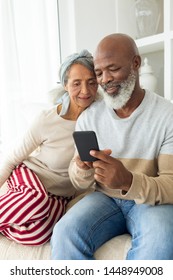 Front View Of Diverse Couple Using A Smartphone On Sofa. Authentic Senior Retired Life Concept