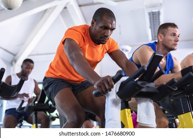 Front View Of Diverse Fit Men Exercising On Exercise Bike In Fitness Center. Bright Modern Gym With Fit Healthy People Working Out And Training At Spin Class