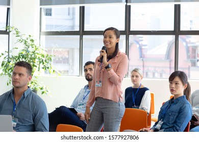 Front view of a diverse group of business creatives sitting in the audience at a business conference, one mixed race young woman standing and using a microphone to ask a question - Powered by Shutterstock