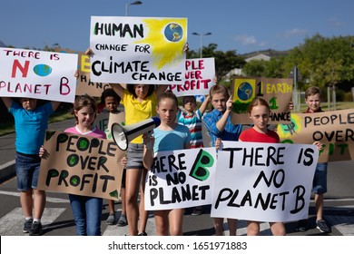 Front view of a diverse group of elementary school pupils walking down a road in the sun on a protest march, carrying signs with environmental and conservation slogans on them, one girl holding a