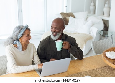 Front view of diverse senior couple using laptop on table while man holds a cup in beach house. Authentic Senior Retired Life Concept - Powered by Shutterstock