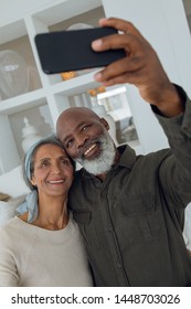 Front view of diverse senior couple taking a picture with smartphone inside a room in beach house. Authentic Senior Retired Life Concept