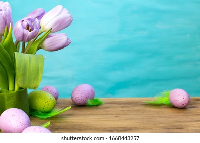 Front View Of A Easter Eggs With Green Feathers, Purple Tulips On Wood And Turquoise Background With Message Space.