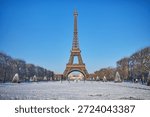 Front view of the Eiffel Tower rising above a snow-covered park in Paris on a sunny winter day. Blue sky, snowy grounds, and trimmed trees create a postcard-perfect scene