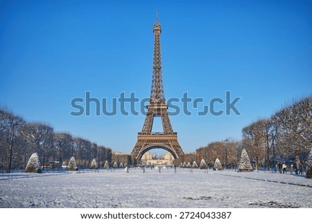 Front view of the Eiffel Tower rising above a snow-covered park in Paris on a sunny winter day. Blue sky, snowy grounds, and trimmed trees create a postcard-perfect scene