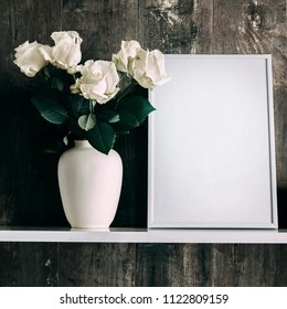 Front view of an empty photo frame layout and a bouquet of white roses in a vase. White flowers on a wooden shelf in the background of vintage wall. square