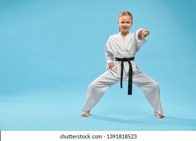 Front view of female teenager standing in stance and punching on blue isolated background. Smiling girl looking at camera and posing in studio. Child fighting and exercising karate. Concept of sport.