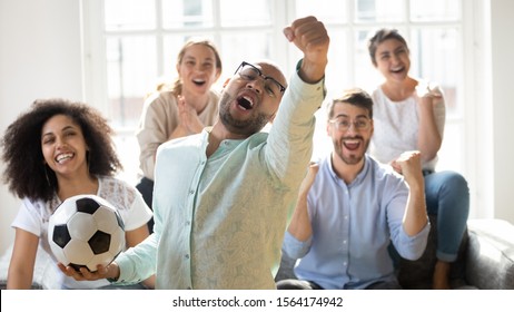 Front View Focus On Overjoyed African American Young Guy Holding Football Ball, Celebrating Victory Of Favorite Team With Friends. Euphoric Mixed Race People Watching Sport Championship On Tv.