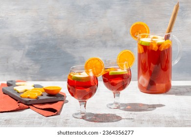 Front view of freshly prepared beautiful red coloured sangria served in glass pitcher on white vintage table, garnished with apples and oranges and in two glasses filled with ice