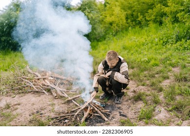 Front view of frozen survivalist male in raincoat putting firewood on campfire to making fire on bank of river in evening before sunset. Concept of exploration, travel and adventure. - Powered by Shutterstock