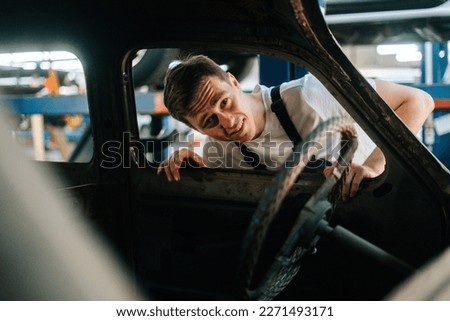 Front view of frustrated young service man in uniform inspecting interior of old car in auto repair shop garage with vehicle background. Concept of car service, repair and maintenance.