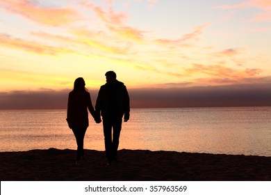 Front view of a full body of couple silhouettes holding hands and walking together looking each other in a date at sunset on the beach