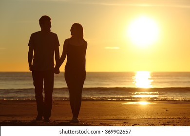 Front view of a full body of a couple silhouette walking together on the beach at sunset in summer