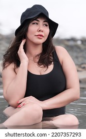 Front View Of Full Figured Young Adult Model In Black One Piece Bathing Swimsuit And Panama Hat Sitting In Outdoors Pool At Spa Resort Having Medicinal Properties. Plus Size Busty Female Looking Away.