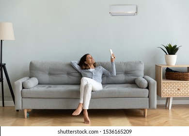 Front view full length joyful young mixed race woman relaxing on cozy couch in living room, holding remote controller, turning on cooler system air conditioner, setting comfortable temperature. - Powered by Shutterstock