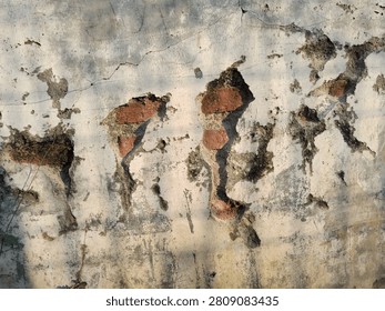 Front view and full-frame shot of an old weathered white concrete wall with peeling plaster, revealing ancient red bricks and rough cement structure underneath
