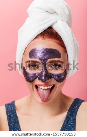 Front view of funny girl showing tongue while doing spa treatment. Studio shot of blissful woman with face mask posing on pink background.