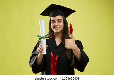 Front View Of Girl In Mortarboard And Graduate Gown Holding Diploma. Pretty Female Graduating From College, University, Showing Super. Isolated On Green Studio Background.