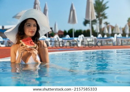 Front view of girl in white swimsuit and hat standing, holding watermelon, eating. Seductive young female standing in pool in water, looking forward, smiling, Concept of youth.