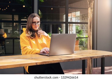 Front View. Girl In Yellow Hoodie Sits In Cafe At Table In Front Of Open Laptop Computer. Woman Works On Computer, Checks Email. Freelancer Works Remotely. Online Education For Adult. Lifestyle.