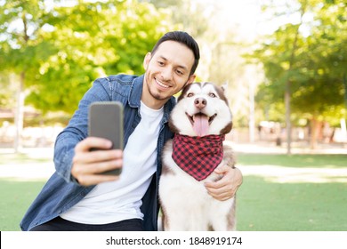 Front view of a good looking man in his 30s taking a photo with his beautiful dog wearing a bandana in the grass at the park