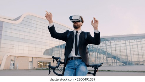 Front view of good-looking concentrated 30-aged bearded man in business clothes which leaning on his bike and working on imaginary screen in virtual reality headset near modern glass building - Powered by Shutterstock