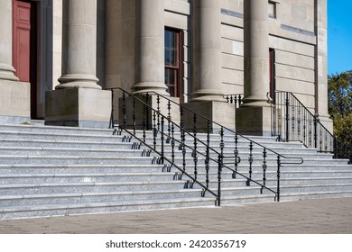 A front view of a government building with large round pillars, red wooden doors, black metal rails, grey marble steps, and a brick entrance. There are trees in the background with a bright blue sky. - Powered by Shutterstock