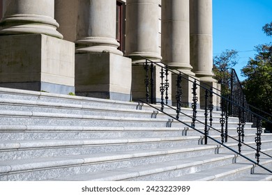 A front view of a government building with large round pillars, red wooden door, black metal rails, grey marble steps, and a brick entrance. There are trees in the background with a bright blue sky. - Powered by Shutterstock