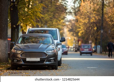 Front view of gray expensive luxurious car parked in quiet alley on sunny autumn day on blurred vehicles, walking people and golden foliage bokeh background. Transportation, parking problems concept.