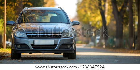 Front view of gray shiny empty car parked in quiet area on wide alley under big trees on blurred green and yellow folliage bokeh background on bright sunny day. Transportation and parking concept.