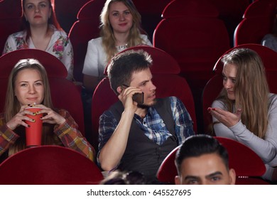 Front View Of Group Of People Watching Film In Cinema Hall, Brunet Man Talking At Call Phone, Angry And Shocked Woman Gesturing By Hand Stopping Him. Leisure And Culture Of Young Girls And Boy.