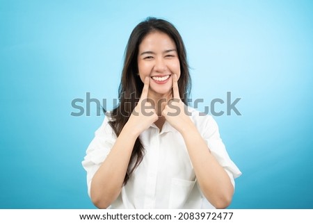 Front view half-body image of a happy Asian woman standing using an index fingers touching the corners of mouth while smiling broadly showing her beautiful white teeth isolated on turquoise background
