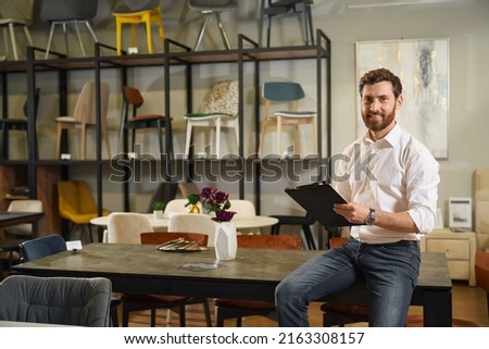 Front view of handsome designer, architect seller sitting on table, writing. Brunette male with beard looking at camera, smiling, designing furniture. Concept of urban lifestyle.