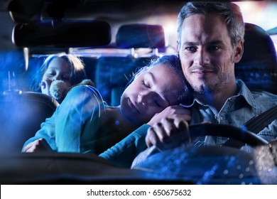 Front View. Handsome Man Driving His Car At Night, His Wife Fell Asleep On His Shoulder. At The Back, His Little Girl Fell Asleep With Her Teddy In Her Arms