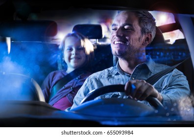 Front View. A Handsome Man Driving His Car At Night In The Rain.  At The Back His Little Daughter Smiles To Her Dad In The Mirror