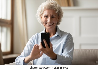 Front View Happy 70s Grandmother Taking Selfie On Smartphone, Using Funny Photo Editing Apps On Mobile Phone. Excited Pleasant Older Woman Reading Good News, Sharing With Friends In Social Network.