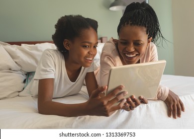 Front view of happy African American mother and daughter lying on bed and using digital tablet in bedroom at home. Social distancing and self isolation in quarantine lockdown for Coronavirus Covid19
