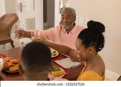 Front view of happy African American grandfather sitting at the dinning table and raising his glass of water for toasting with his family at home
