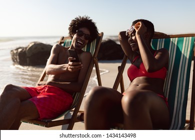 Front view of happy African-american couple having fun on mobile phone while relaxing in a beach chair on the beach