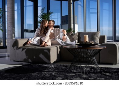 Front View Of Happy Couple Lying On Sofa And Hugging Near Large Panoramic Window With Incredible View On City. Concept Of Enjoying Free Time At Home Together.