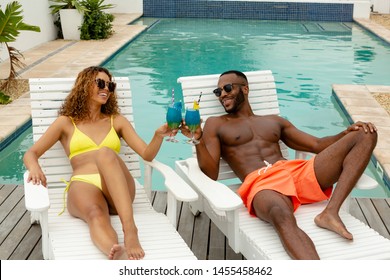 Front view of happy diverse couple toasting glasses of cocktail while relaxing on a sun lounger near swimming pool. Summer fun at home by the swimming pool - Powered by Shutterstock