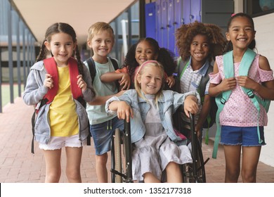 Vista frontal de colegiales diversos y felices parados en el pasillo exterior del colegio mientras una colegiala caucásica está sentada en silla de ruedas en primer plano