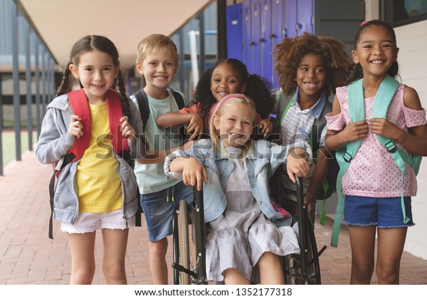 学校の廊下の外に立つ幸せな多様な学校の子どもたちの正面図 前景に白人の女学生が車椅子に座っている の写真素材 今すぐ編集