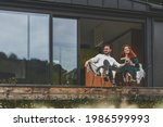 Front view of happy family sitting outdoor on the terrace of country house. Stylish young couple on weekend sits and enjoys the nature view on the terrace of their modern home with panoramic windows.