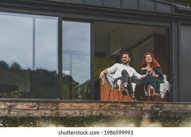 Front View Of Happy Family Sitting Outdoor On The Terrace Of Country House. Stylish Young Couple On Weekend Sits And Enjoys The Nature View On The Terrace Of Their Modern Home With Panoramic Windows.